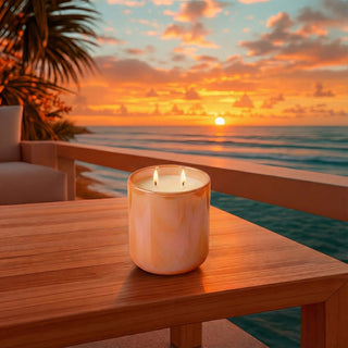 Sun-Kissed Glow – Staycation sunset-inspired premium coconut apricot wax candle lit with dual warm flames, radiant iridescent peach glass vessel on wooden deck overlooking ocean waves & vibrant golden hour sunset sky, ultimate tropical escape ambiance