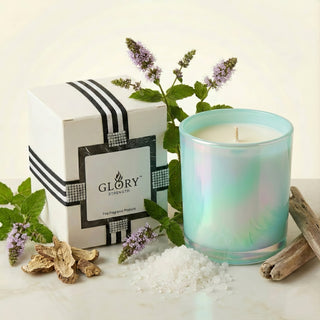 Glory Strength Shimmering Sea 8oz luxury candle in an iridescent turquoise vessel and white gift box, surrounded by fresh mint botanicals, dried driftwood, sea salt crystals, and dried roots representing the candle's ocean-inspired fragrance notes.