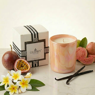 Glory Strength Sun-Kissed Glow 8oz luxury candle in an iridescent peach vessel and white gift box, surrounded by fresh passionfruit, guava, frangipani flowers, and vanilla beans representing the candle's tropical sunset-inspired fragrance notes.