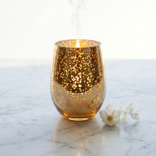 Glory Strength Tomorrow candle from the Legacy Collection — antique gold mercury glass vessel lit on white Carrara marble with white magnolia blooms, gossamer smoke trail rising against a bright open background
